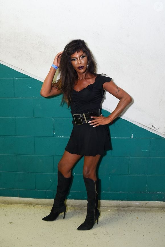 Max Weber: exibe um visual dark glam arrojado com um vestido curto preto de decote com leve franzido e alças delicadas, marcado por um cinto largo de couro com fivela retangular imponente na cintura. A composição é finalizada por botas pretas de camurça de salto fino e cano alto, maquiagem social marcante com lábios escuros e cabelos volumosos desfiados.