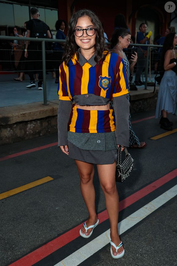 Samantha Alves desfilou uma estética preppy desconstruída e vibrante, utilizando um conjunto de tricô listrado em azul, amarelo e bordô, que remete a uniformes escolares clássicos com um toque subversivo. O look apresenta um corte cropped tanto na blusa quanto na sobreposição, revelando uma base de alfaiataria cinza risca-de-giz por baixo