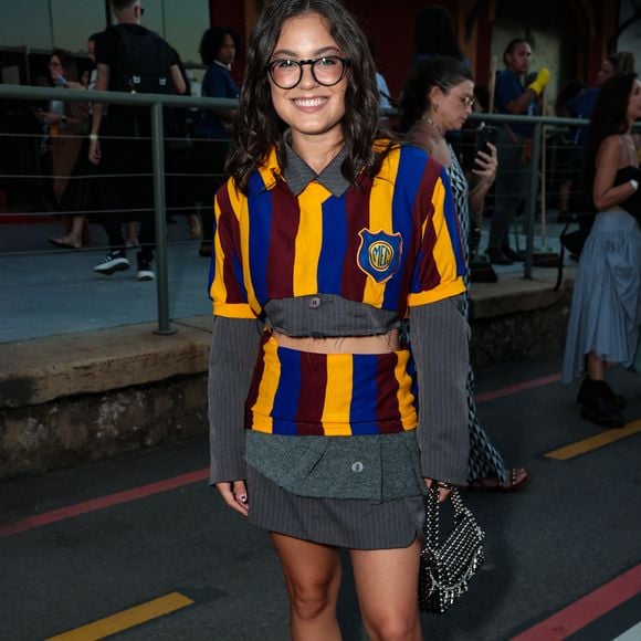 Samantha Alves desfilou uma estética preppy desconstruída e vibrante, utilizando um conjunto de tricô listrado em azul, amarelo e bordô, que remete a uniformes escolares clássicos com um toque subversivo. O look apresenta um corte cropped tanto na blusa quanto na sobreposição, revelando uma base de alfaiataria cinza risca-de-giz por baixo