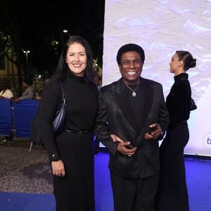 Neguinho da Beija-Flor e Elaine Reis: O cantor usa um terno preto texturizado com lapela acetinada clássica, camisa preta canelada, cordão com medalha dourada e tênis casual branco com cadarços pretos. Sua esposa veste um vestido preto de gola alta, cinto fino, botas de cano alto e bolsa de ombro azul-marinho metalizada