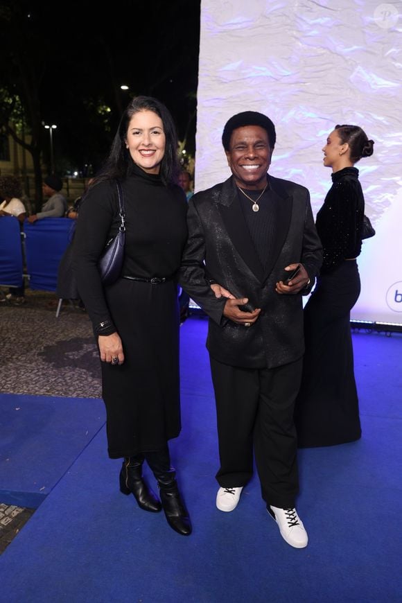 Neguinho da Beija-Flor e Elaine Reis: O cantor usa um terno preto texturizado com lapela acetinada clássica, camisa preta canelada, cordão com medalha dourada e tênis casual branco com cadarços pretos. Sua esposa veste um vestido preto de gola alta, cinto fino, botas de cano alto e bolsa de ombro azul-marinho metalizada
