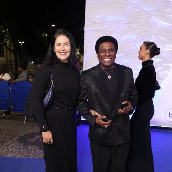 Neguinho da Beija-Flor e Elaine Reis: O cantor usa um terno preto texturizado com lapela acetinada clássica, camisa preta canelada, cordão com medalha dourada e tênis casual branco com cadarços pretos. Sua esposa veste um vestido preto de gola alta, cinto fino, botas de cano alto e bolsa de ombro azul-marinho metalizada