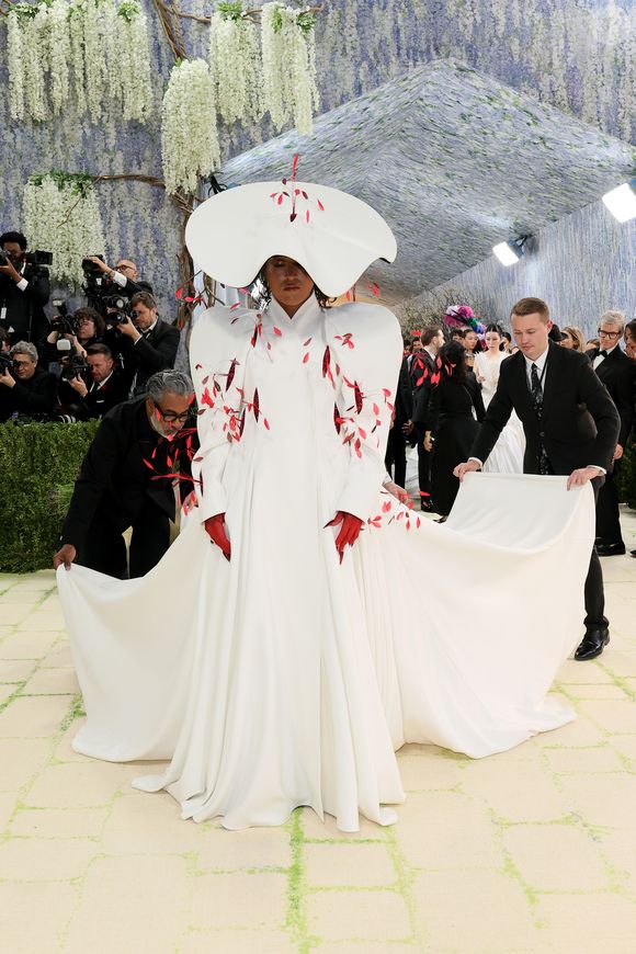 Naomi Osaka apostou na dramaticidade com um volumoso vestido branco de gola alta e ombreiras arquitetônicas, adornado com aplicações que remetem a pétalas vermelhas, finalizando com um chapéu monumental em formato de disco