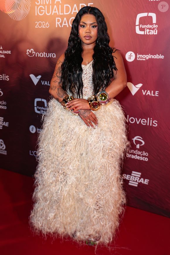 A cantora Duquesa brilhou em um vestido off-white que brinca com texturas volumosas e detalhes minuciosos. A peça apresenta um corpete em crochê ou renda delicada, que se transforma em uma saia ampla coberta por franjas longas e macias, criando um efeito de movimento impactante