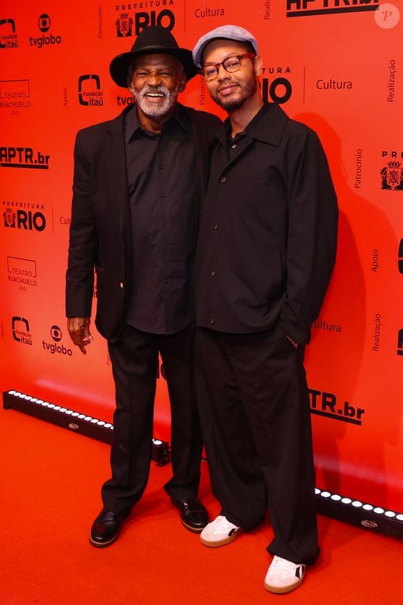 Antônio Pitanga e Bento Ribeiro: Antônio Pitanga veste um terno preto clássico de corte reto alinhado a uma camisa social também preta, arrematando a elegância do visual com sapatos sociais escuros e um chapéu fedora preto. Bento Ribeiro opta por uma linha casual contemporânea toda em preto, composta por uma camisa de botões de caimento solto, calça oversized e tênis brancos com detalhes escuros, finalizando a produção com uma boina cinza claro