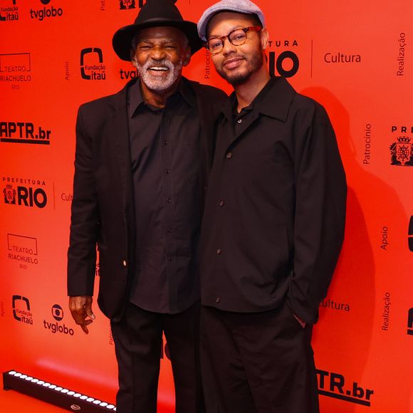 Antônio Pitanga e Bento Ribeiro: Antônio Pitanga veste um terno preto clássico de corte reto alinhado a uma camisa social também preta, arrematando a elegância do visual com sapatos sociais escuros e um chapéu fedora preto. Bento Ribeiro opta por uma linha casual contemporânea toda em preto, composta por uma camisa de botões de caimento solto, calça oversized e tênis brancos com detalhes escuros, finalizando a produção com uma boina cinza claro