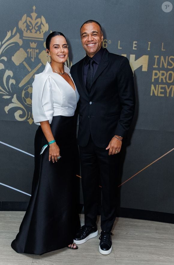 Denílson e Luciele di Camargo: Luciele vestiu um modelo bicolor em branco e preto com decote V profundo, mangas estruturadas e saia longa fluida; Denílson optou por um terno monocromático preto com camisa e gravata na mesma cor, finalizado por tênis de sola branca