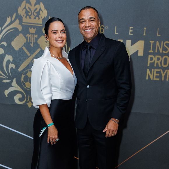 Denílson e Luciele di Camargo: Luciele vestiu um modelo bicolor em branco e preto com decote V profundo, mangas estruturadas e saia longa fluida; Denílson optou por um terno monocromático preto com camisa e gravata na mesma cor, finalizado por tênis de sola branca