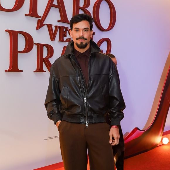 O ex-BBB Vinicius investiu em uma paleta de tons terrosos e texturas contrastantes. Ele veste uma jaqueta bomber de couro preto oversized com fechamento em zíper, sobreposta a uma camisa polo em tom de marrom café. A calça de alfaiataria possui corte reto e modelagem ampla (wide leg) em um tom de marrom médio