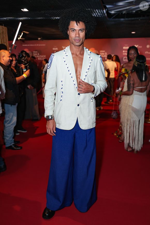 Breno Simões marcou presença com um visual que equilibra alfaiataria clássica e elementos náuticos. O ex-BBB veste um blazer off-white de linho, usado sem camisa, que se destaca pelos detalhes de búzios e cordões azuis aplicados na lapela e nos ombros
