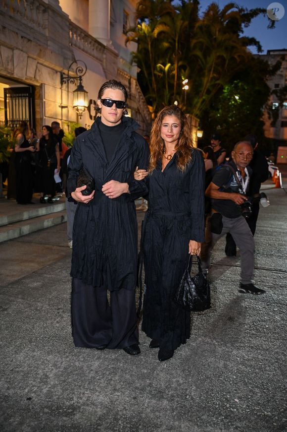 Isadora Cruz e Lucas Pinheiro: o casal surgiu em uma proposta dark academia fundida ao utilitarismo moderno, apostando no impacto visual das texturas monocromáticas. Isadora Cruz veste um macacão preto de tecido anasalado e aspecto 'amassado' proposital, com amarração na cintura e decote em "V", complementado por uma bolsa de ombro no mesmo material e um colar de elos dourados que ilumina o rosto
