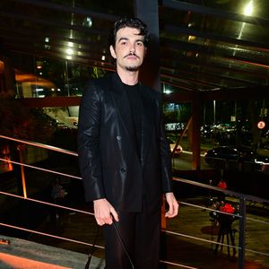 Jão: o cantor veste um terno preto em tom acetinado, composto por blazer trespassado (double-breasted) de abotoamento duplo e calça reta fluida. Por baixo, utiliza uma camiseta preta básica de gola careca. Como acessório, carrega uma bolsa de ombro em couro marrom-escuro estruturada e calça sapatos pretos de bico fino