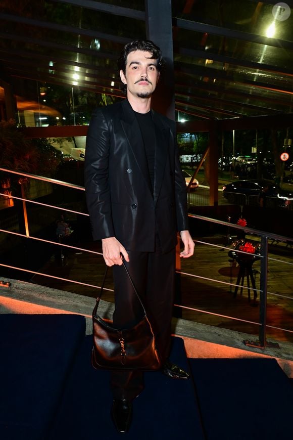 Jão: o cantor veste um terno preto em tom acetinado, composto por blazer trespassado (double-breasted) de abotoamento duplo e calça reta fluida. Por baixo, utiliza uma camiseta preta básica de gola careca. Como acessório, carrega uma bolsa de ombro em couro marrom-escuro estruturada e calça sapatos pretos de bico fino