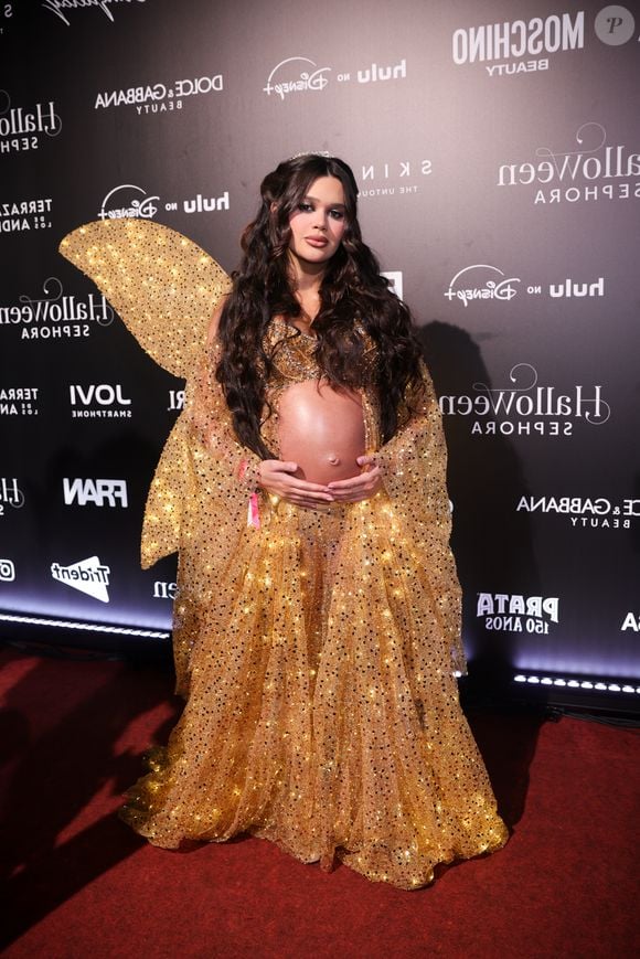 Franciny Ehlke, de 'guardiã da luz', veste um conjunto dourado reluzente de inspiração de fada, composto por top cropped estruturado e saia longa rodada com transparência e aplicação de lantejoulas, deixando a barriga de grávida à mostra. A produção conta com mangas amplas e fluídas do mesmo tecido brilhante, asas douradas estruturadas nas costas, cabelos ondulados soltos com tiara discreta e maquiagem em tons terrosos iluminados