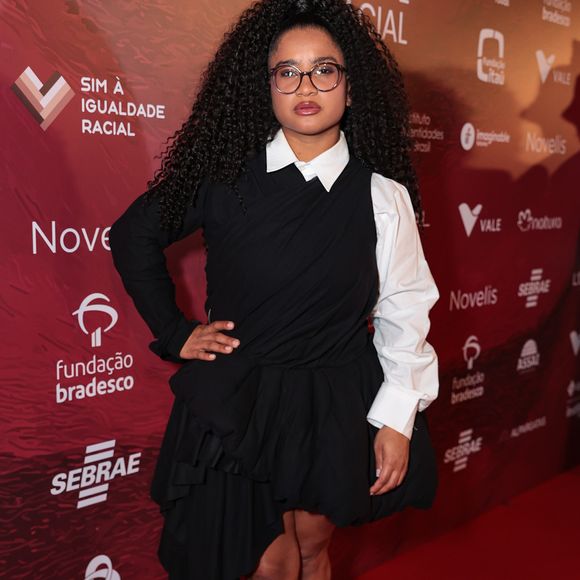 Tia Milena (de cabelão!) optou por uma proposta desconstruída e contemporânea, utilizando um vestido preto de modelagem assimétrica com drapeados volumosos na saia. A peça é sobreposta a uma camisa branca clássica, cujas mangas bufantes e gola estruturada criam um contraste marcante entre o formal e o artístico. Para completar a produção, ela escolheu botas de bico fino em couro preto e óculos de armação discreta, mantendo o foco no jogo de formas do vestuário