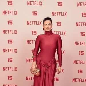 Emanuelle Araújo: veste um vestido longo em tom vermelho-fechado com gola alta e mangas compridas, destacando-se pelo nó drapeado na cintura com caimento fluido na saia, acompanhado de clutch dourada e brincos combinando