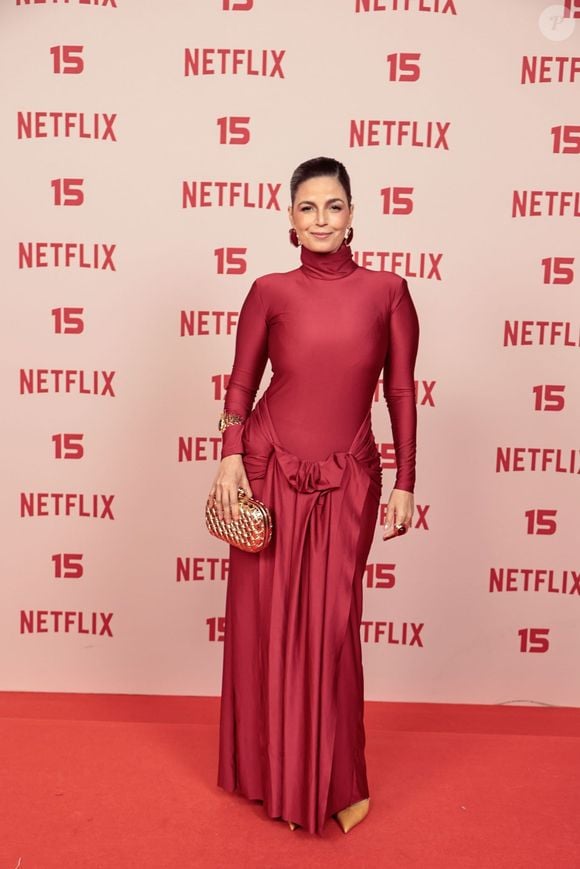 Emanuelle Araújo: veste um vestido longo em tom vermelho-fechado com gola alta e mangas compridas, destacando-se pelo nó drapeado na cintura com caimento fluido na saia, acompanhado de clutch dourada e brincos combinando