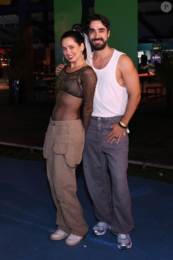 Raissa Xavier e Gabriel Godoy: Raissa investiu no visual streetwear ousado com uma blusa de tule preta com brilhos dourados totalmente transparente sobre um top escuro, combinada com calça cargo bege de bolsos utilitários volumosos e tênis brancos. Gabriel acompanhou a estética urbana vestindo regata branca tradicional, calça jeans cinza de corte solto e tênis esportivo metalizado em tons de azul e prata