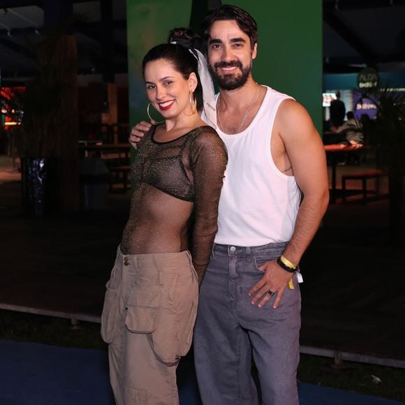 Raissa Xavier e Gabriel Godoy: Raissa investiu no visual streetwear ousado com uma blusa de tule preta com brilhos dourados totalmente transparente sobre um top escuro, combinada com calça cargo bege de bolsos utilitários volumosos e tênis brancos. Gabriel acompanhou a estética urbana vestindo regata branca tradicional, calça jeans cinza de corte solto e tênis esportivo metalizado em tons de azul e prata