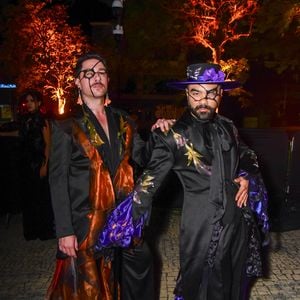 Mauro Souza e Rafael Piccin vestem trajes temáticos em preto e cores vibrantes. O primeiro (à esquerda) veste um conjunto preto com lapelas e babados volumosos na saia em tecido transparente laranja acobreado. Ele usa um tapa-olho preto com detalhes e maquiagem dramática nos olhos. O segundo (à direita) veste um traje preto com lapelas pretas e babados roxos vibrantes nas mangas e na barra da calça, além de um lenço de pescoço roxo e um chapéu roxo com aba preta decorado com uma flor roxa e uma rede preta sobre o rosto. Ambos usam maquiagem facial preta