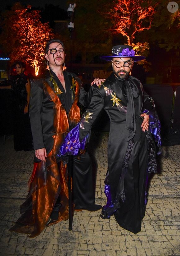 Mauro Souza e Rafael Piccin vestem trajes temáticos em preto e cores vibrantes. O primeiro (à esquerda) veste um conjunto preto com lapelas e babados volumosos na saia em tecido transparente laranja acobreado. Ele usa um tapa-olho preto com detalhes e maquiagem dramática nos olhos. O segundo (à direita) veste um traje preto com lapelas pretas e babados roxos vibrantes nas mangas e na barra da calça, além de um lenço de pescoço roxo e um chapéu roxo com aba preta decorado com uma flor roxa e uma rede preta sobre o rosto. Ambos usam maquiagem facial preta