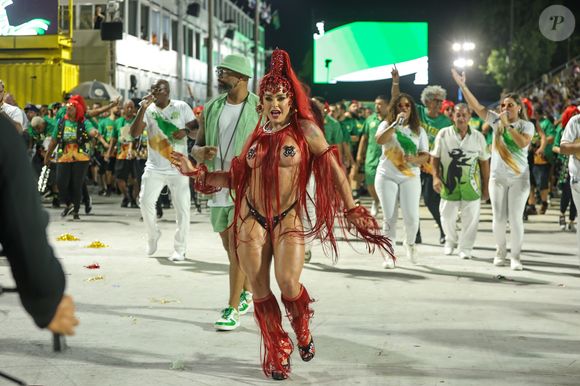 Os fios escarlates caem do cocar até abaixo da linha dos quadris, criando movimento constante e cobrindo parcialmente o corpo. O adereço de cabeça é adornado com pedrarias vermelhas e cristais, formando uma espécie de máscara frontal que emoldura o rosto