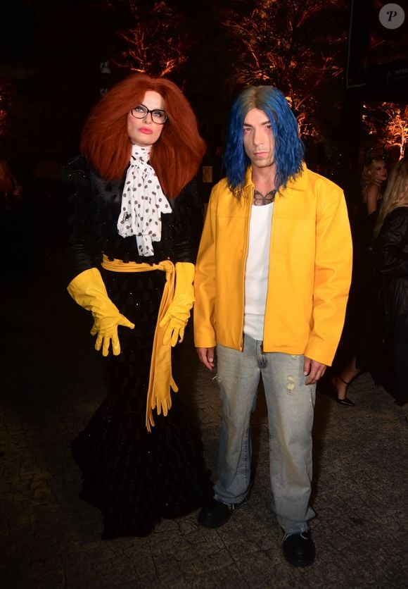Di Ferrero e Isabeli Fontana: à esquerda, o figurino traz um vestido longo preto sereia com textura pontilhada, cinto largo amarelo amarrado na cintura e luvas amarelas de cano longo. O visual inclui lenço de gola branco com bolinhas pretas, óculos de armação preta, batom rosa e uma peruca volumosa ruiva. À direita, o look casual é composto por jaqueta de couro sintético amarela, camisa branca básica, calça jeans clara de modelagem reta e sapatos pretos. A caracterização conta com peruca azul lisa de corte médio e pintura/tatuagem no peito