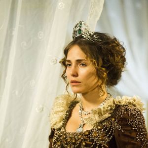 Leticia Colin em 'Novo Mundo': penteado de época sobre base de cabelo longo e cacheado. O topo é preso e polido, enquanto as laterais e o comprimento apresentam cachos definidos e repicados para volume. A cor é um castanho escuro com luzes acobreadas sutis nos cachos frontais