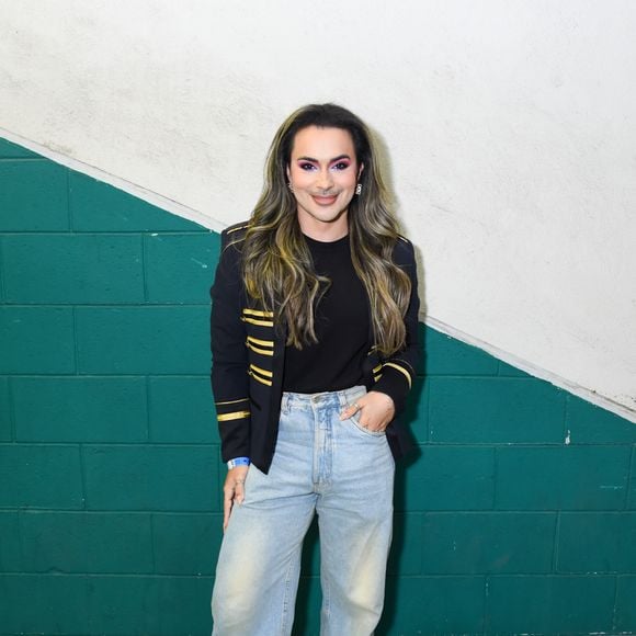 Andy Rodriguez: aposta em um estilo casual chic com inspiração militar, vestindo um blazer preto estruturado com detalhes de listras douradas nas mangas e nos ombros. A peça sobrepõe uma camiseta preta lisa, combinada com calça jeans clara de modelagem ampla, botas pretas de solado tratorado e uma maquiagem marcante com sombra em tons de rosa e roxo