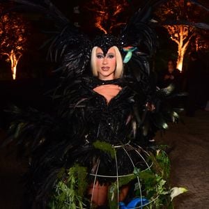 Nathalia Valente veste um macacão justo preto coberto inteiramente por penas pretas volumosas, simulando asas abertas atrás dela e detalhes nos ombros e braços. A peça tem recortes no busto e na cintura. Ela usa uma peruca loira curta e reta e um adereço de cabeça que lembra uma cabeça de pássaro com um bico preto falso sobre a testa e duas pequenas réplicas de araras coloridas nas laterais. Nos pés, ela calça botas pretas de plataforma