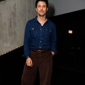 Romulo Estrela apostou em um visual casual elegante com camisa jeans de lavagem escura e botões frontais abertos, combinada a uma calça de veludo cotelê marrom-escuro de modelagem reta. O look é ajustado por um cinto de couro com fivela oval dourada e finalizado com sapatos escuros de sola tratorada