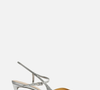 O modelo traz tira slingback cruzada, cabedal bicolor com recorte em "V" sobre a parte superior do pé, ponta fina e salto médio fino de 6,6 centímetros. A construção inclui uma tira delicada que percorre a lateral, envolve o calcanhar, cruza o peito do pé e fecha com fivela metálica. A palmilha é bege e leva a inscrição da marca