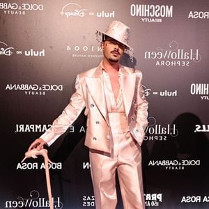 Pedro Sampaio veste um terno de cetim rosa claro cintilante com lapelas largas e botões decorativos. Ele complementa o look com uma camisa de seda rosa claro aberta no peito, uma faixa de cintura larga combinando e calças de corte reto. Ele usa sapatos brancos pontudos e um chapéu de cowboy rosa decorado com strass e franjas de strass, além de segurar uma bengala rosa ornamentada com uma figura de animal na alça