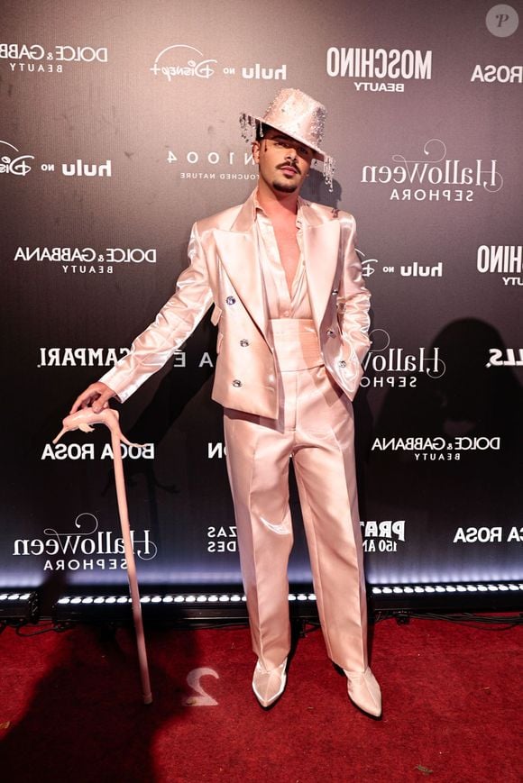 Pedro Sampaio veste um terno de cetim rosa claro cintilante com lapelas largas e botões decorativos. Ele complementa o look com uma camisa de seda rosa claro aberta no peito, uma faixa de cintura larga combinando e calças de corte reto. Ele usa sapatos brancos pontudos e um chapéu de cowboy rosa decorado com strass e franjas de strass, além de segurar uma bengala rosa ornamentada com uma figura de animal na alça