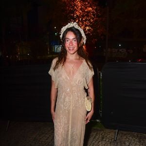Valentina Bandeira veste um vestido longo e fluido em tom bege/marfim com transparência delicada e bordados em pedrarias cintilantes por toda a peça. O visual de estilo ninfa ou fada é arrematado por uma coroa de flores brancas na cabeça, maquilhagem leve com blush rosado destacado nas bochechas e sapatos combinando