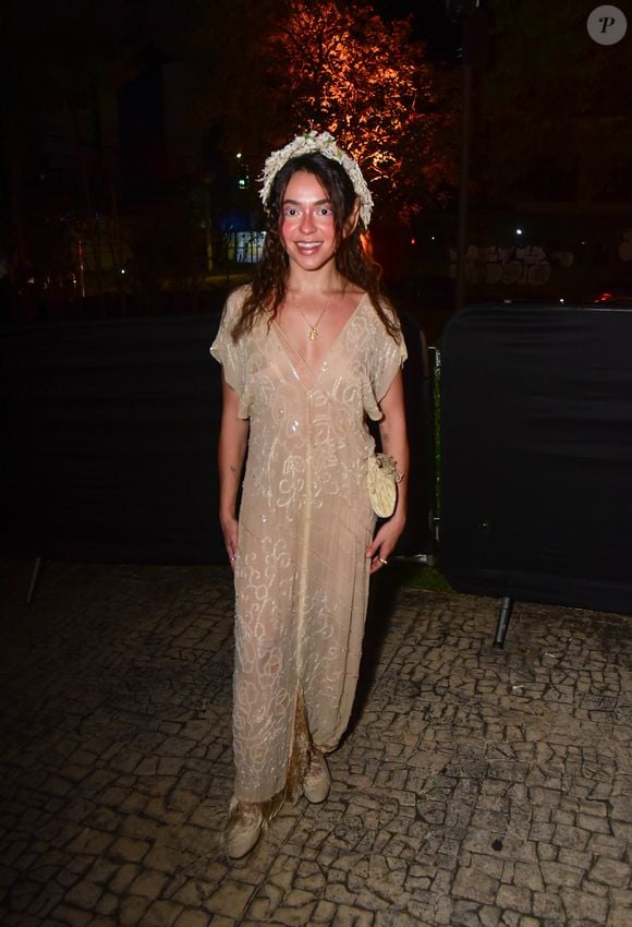 Valentina Bandeira veste um vestido longo e fluido em tom bege/marfim com transparência delicada e bordados em pedrarias cintilantes por toda a peça. O visual de estilo ninfa ou fada é arrematado por uma coroa de flores brancas na cabeça, maquilhagem leve com blush rosado destacado nas bochechas e sapatos combinando