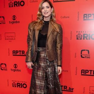 Mel Lisboa exibe um conjunto contemporâneo combinando um blazer de sarja bronze com textura acetinada sobre uma blusa preta transparente de gola alta. A saia midi plissada apresenta um padrão metalizado geométrico, completada por meia-calça escura e sapatos pretos tratorados