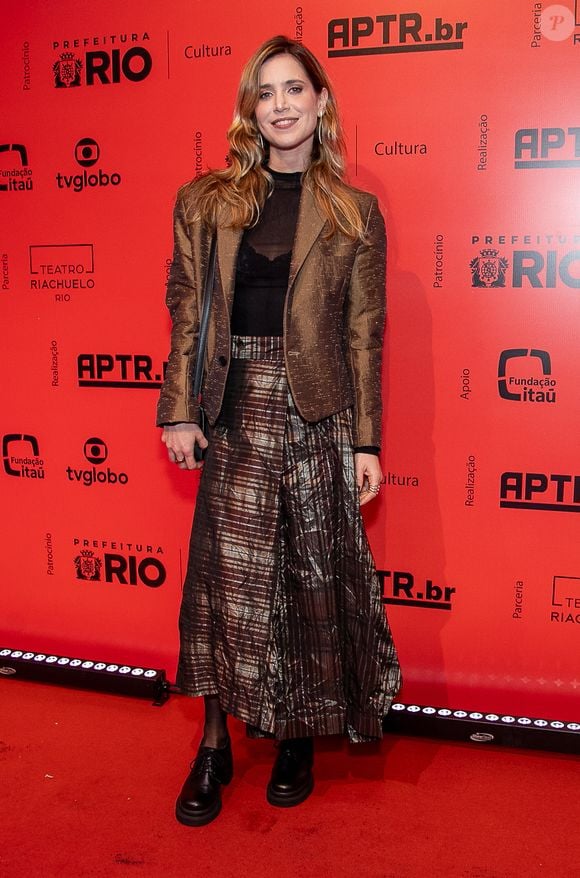 Mel Lisboa exibe um conjunto contemporâneo combinando um blazer de sarja bronze com textura acetinada sobre uma blusa preta transparente de gola alta. A saia midi plissada apresenta um padrão metalizado geométrico, completada por meia-calça escura e sapatos pretos tratorados