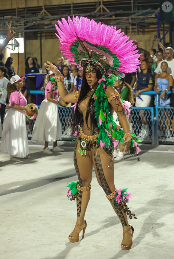 O grande destaque é o cocar circular de plumas rosa, aberto como um sol, com base verde e aplicações de contas e sementes. O adereço cria uma moldura dramática para o rosto e amplia a silhueta horizontalmente