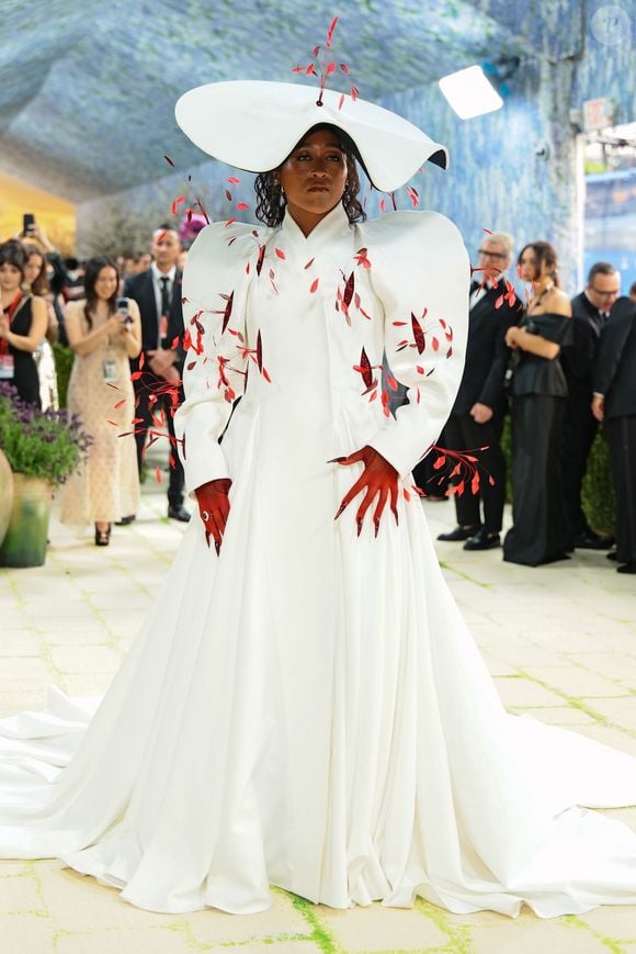 A tenista japonesa Naomi Osaka entregou drama no Met Gala 2026 com um look branco look de Robert Wun com ombros bufantes, chapéu coordenado e plumas vermelhas que contrastaram com a luva da mesma cor