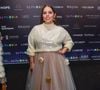 Tati Machado esolheu uma produção que mistura o conforto do tricô com a fantasia do tule. A apresentadora utiliza um suéter de gola alta em tom creme com aplicações de lantejoulas transparentes, criando um brilho molhado sob as luzes. A peça é combinada com uma saia volumosa de tule bege, que ganha um toque 'diferentão' com a aplicação esparsa de flores tridimensionais em tons de branco e amarelo queimado, finalizada com uma maquiagem focada em tons de roxo nos olhos