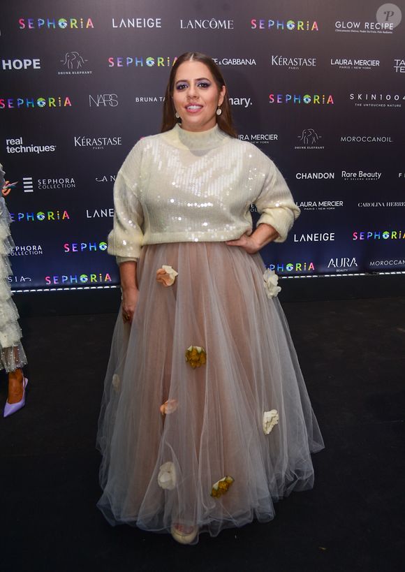 Tati Machado esolheu uma produção que mistura o conforto do tricô com a fantasia do tule. A apresentadora utiliza um suéter de gola alta em tom creme com aplicações de lantejoulas transparentes, criando um brilho molhado sob as luzes. A peça é combinada com uma saia volumosa de tule bege, que ganha um toque 'diferentão' com a aplicação esparsa de flores tridimensionais em tons de branco e amarelo queimado, finalizada com uma maquiagem focada em tons de roxo nos olhos