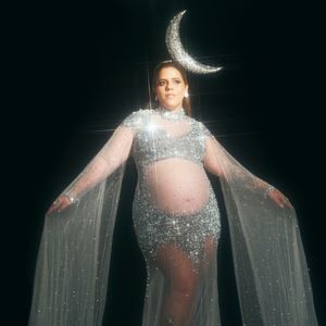 Tati Machado chamou a fantasia de 'Mother Moon': ela veste um vestido transparente prateado coberto de pedrarias e brilhos, desenhado com decote alto e mangas longas em estilo capa que se estendem até o chão. O modelo destaca a barriga de gravidez e conta com fenda sutil, sapatos de salto prateados e uma tiara metálica em formato de meia-lua prateada no topo da cabeça