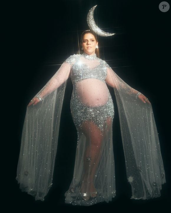 Tati Machado chamou a fantasia de 'Mother Moon': ela veste um vestido transparente prateado coberto de pedrarias e brilhos, desenhado com decote alto e mangas longas em estilo capa que se estendem até o chão. O modelo destaca a barriga de gravidez e conta com fenda sutil, sapatos de salto prateados e uma tiara metálica em formato de meia-lua prateada no topo da cabeça