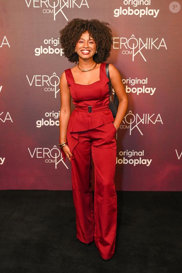 Emmy Neves: a atriz escolheu um macacão vermelho-fechado sem mangas, com decote reto estruturado e detalhe de peplum assimétrico na cintura. A peça é acinturada por um cinto de tecido do próprio macacão com fivela metálica escura e possui calça de corte reto amplo. Acessoriza o look com gargantilha rígida dourada, pulseiras combinando e bolsa preta de ombro