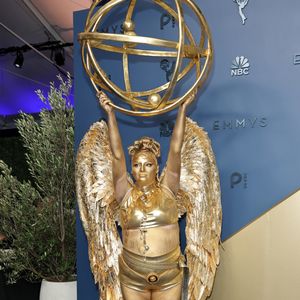 Megan Stalter: apostando em uma caracterização performática e literal da própria estatueta do Emmy, o look monocromático em tom dourado reluzente traz top cropped e hot pants metálicos complementados por imponentes asas de penas esculpidas nas costas. Toda a pele e o cabelo aparecem cobertos por tinta corporal metálica no mesmo tom, emoldurados por colares e cinto de correntes