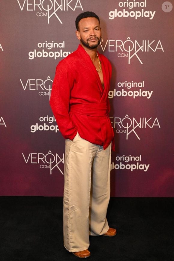 Dhério Chagas: o ator veste um terno desconstruído em kimono vermelho-fechado, com tecido de textura leve, mangas longas e transpassado ajustado na cintura por amarração. A peça deixa o tórax parcialmente à mostra, onde ostenta uma corrente dourada espessa. Na parte inferior, utiliza calça alfaiataria de pernas largas em tom bege-areia e mocassins de couro marrom