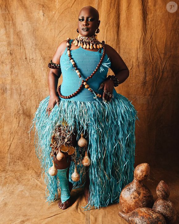 Rob Freitas, fazendo alusão à Oxossi, veste um top regata azul estruturado combinado com uma saia volumosa confeccionada em tiras de ráfia ou palha azul. O visual étnico-artístico é enriquecido por múltiplos colares e colares transversais de contas e sementes de madeira, maxi gargantilha com búzios e pedrarias, braçadeiras de contas e enfeites pendentes de porongos/cabaças na saia e na perna