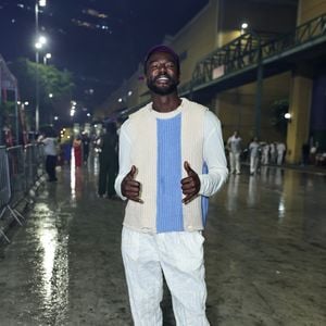 Jonathan Azevedo vestiu uma composição sobreposta composta por camiseta de mangas compridas branca sob um colete de tricô estruturado nas cores bege e azul central. Na parte inferior, usou calça ampla clara com textura levemente estampada/lavada e tênis em tom bege com detalhes azuis. O visual foi arrematado com um boné roxo usado virado para trás