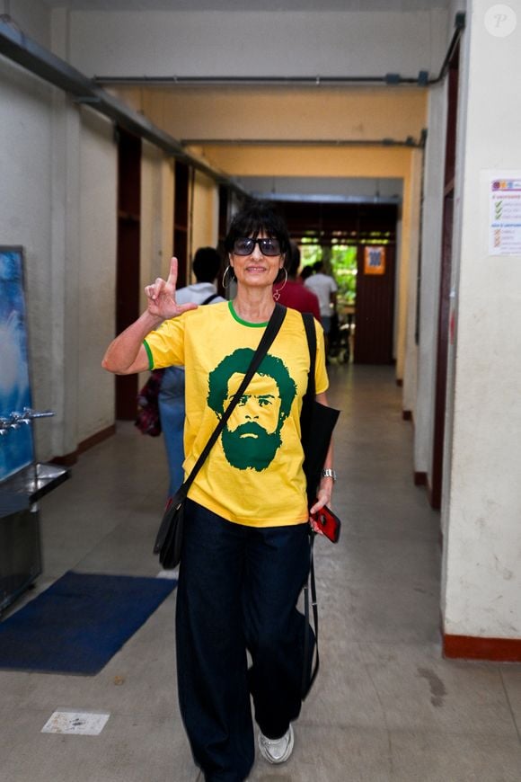 Fernanda Abreu veste blusa nas cores do Brasil com o rosto de Lula e faz o 'L' para fotógrafos em colégio eleitoral